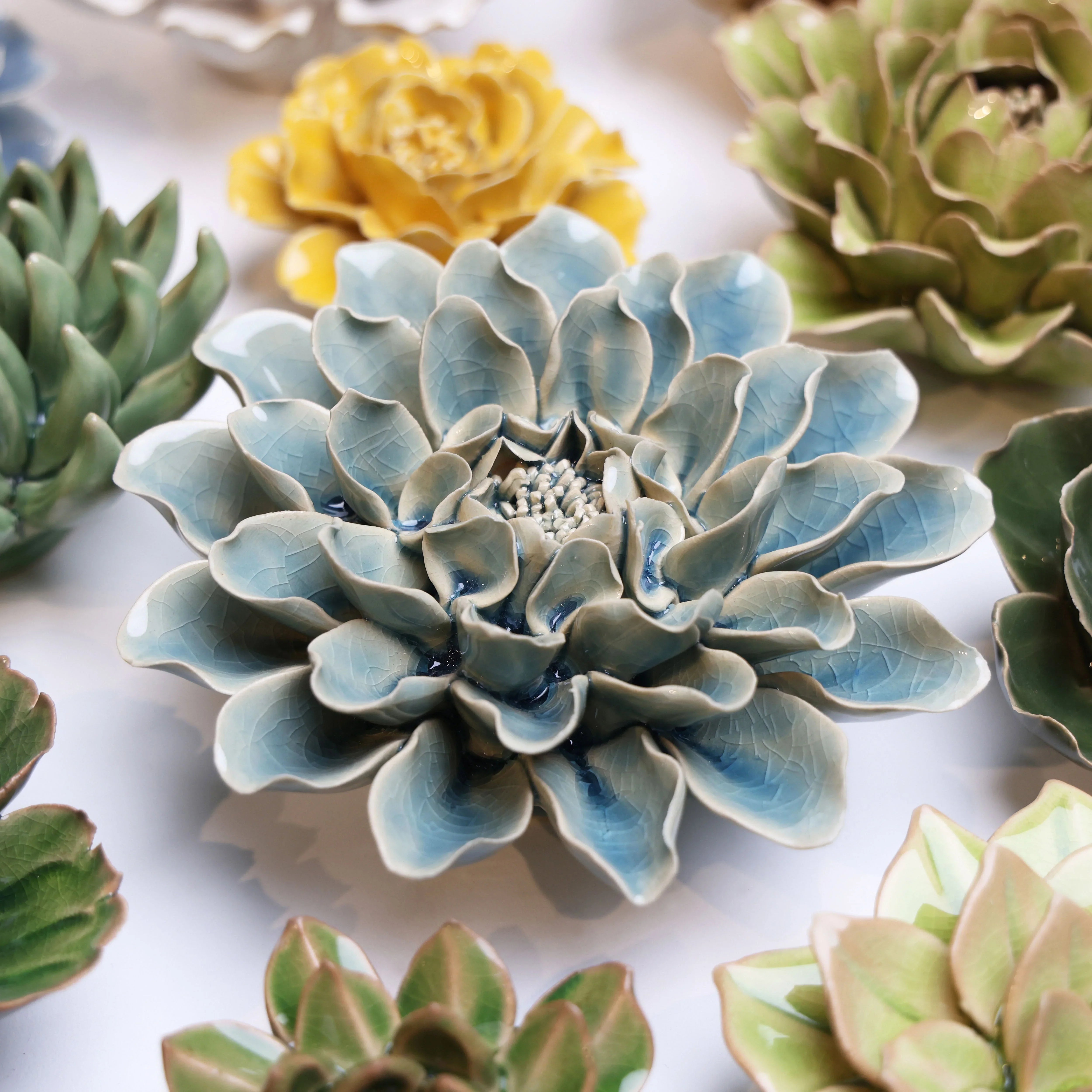 A group of ceramic flowers in various colors, including blue, green, and yellow, are arranged on a white surface. The flowers' textured petals create a lifelike, delicate appearance.