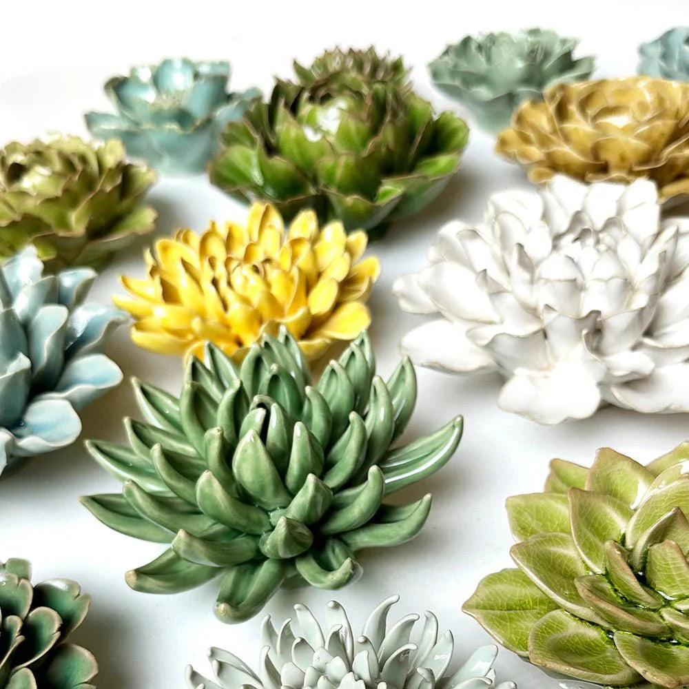 Colorful ceramic flowers arranged on a white background. The centerpiece is a large pink flower, surrounded by green, blue, purple, and yellow ceramic flowers with detailed petals and leaves.