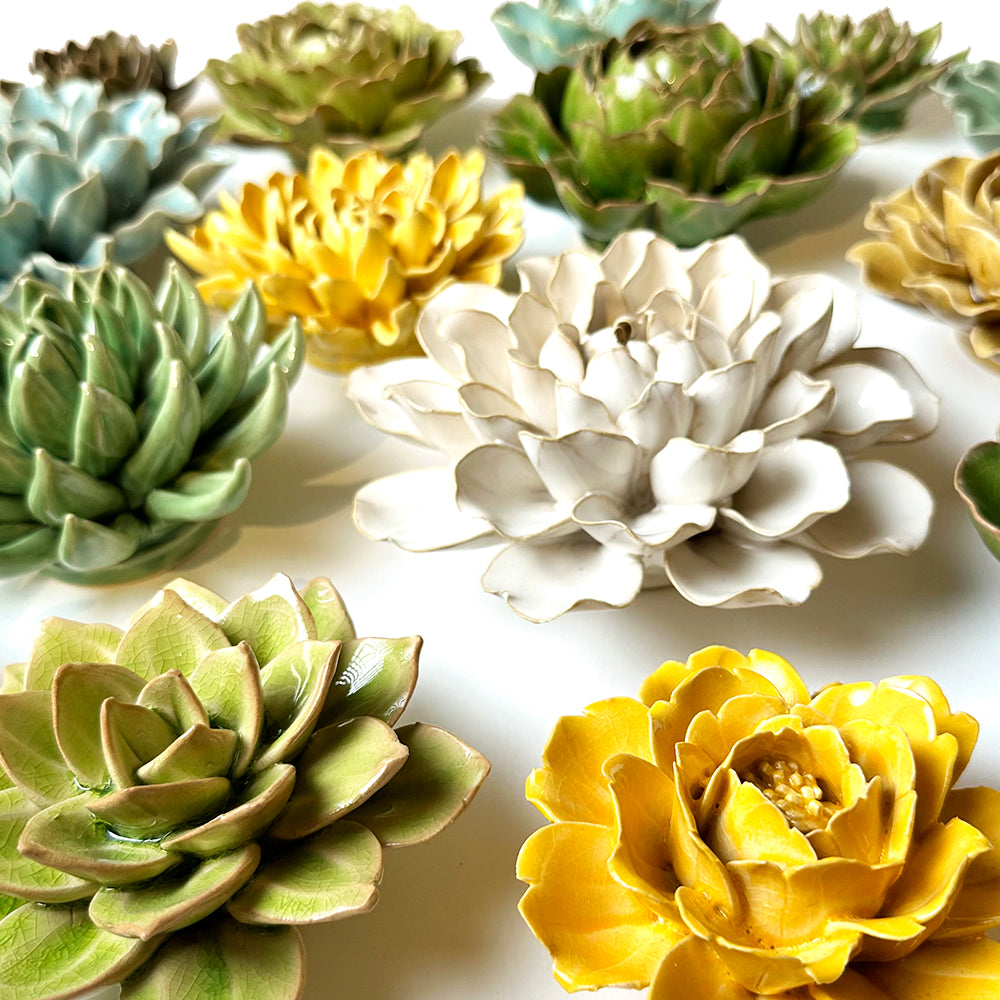 Ceramic wall flowers in shades of yellow, green, white, and blue are arranged on a white background. Each piece is detailed, resembling real flower patterns.