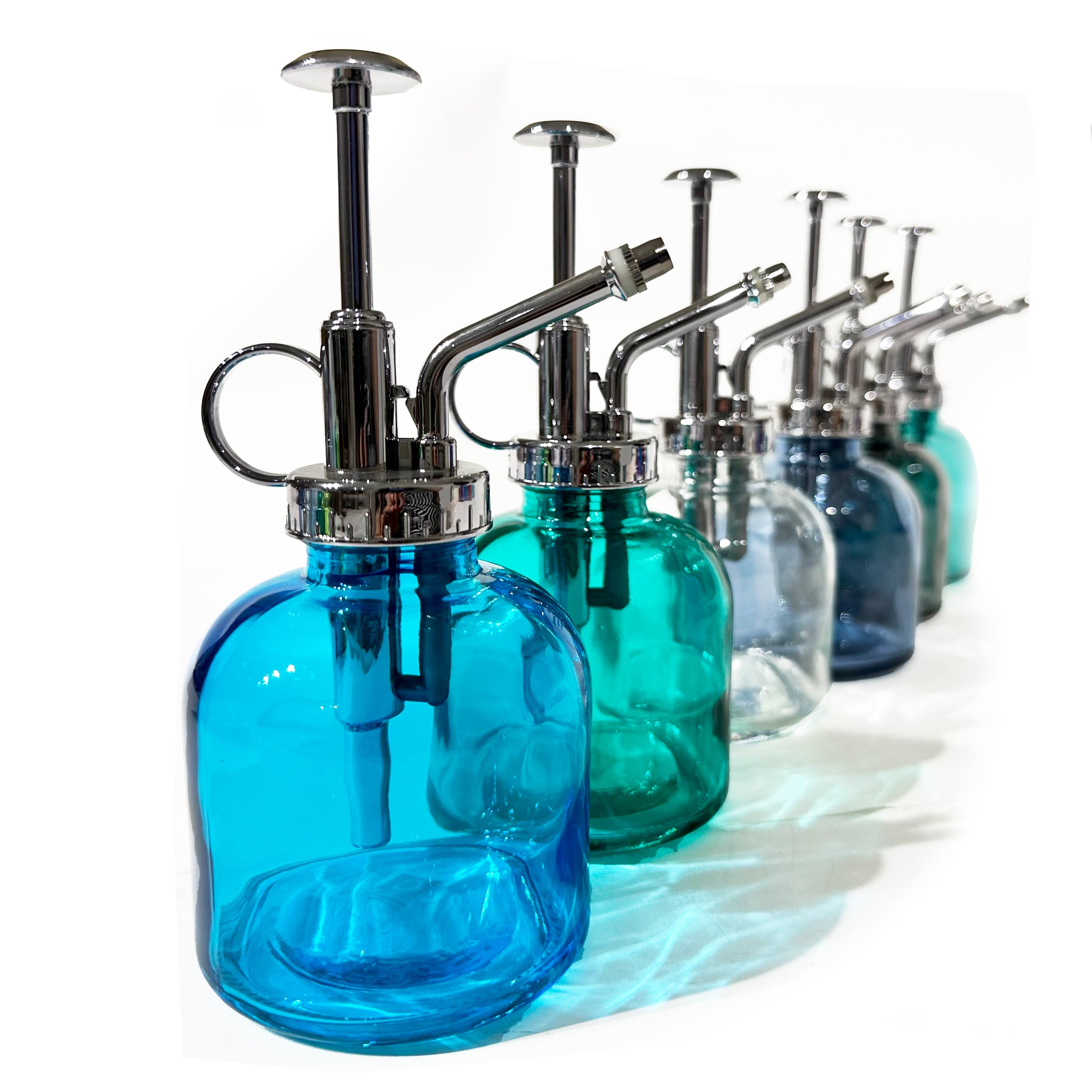 A row of colorful glass plant misters in shades of blue, green, and clear, with silver metal pumps, arranged on a white surface.