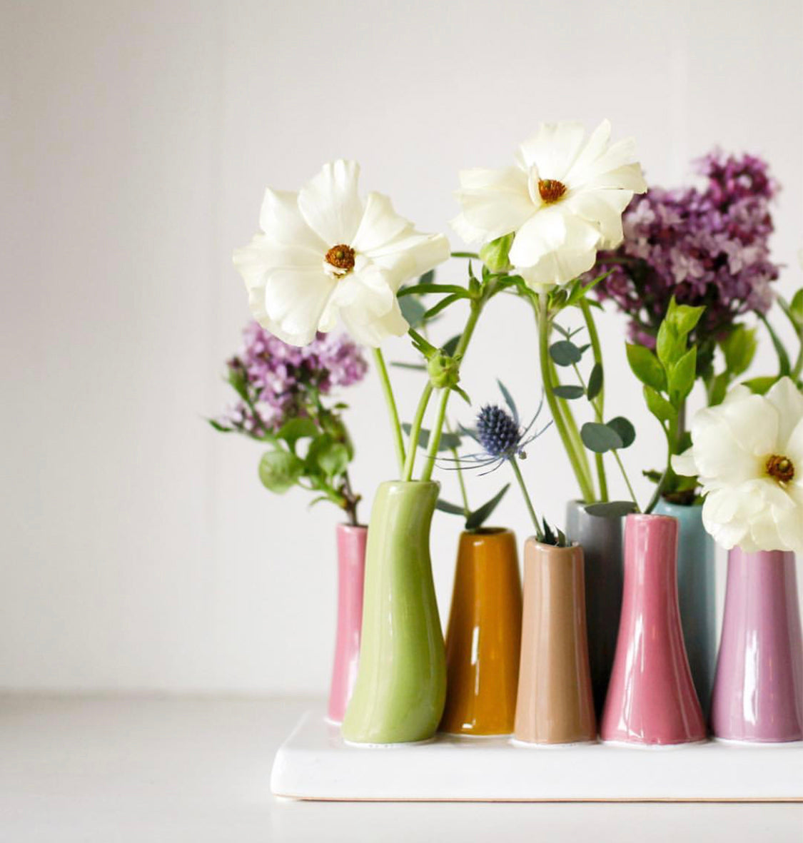 A row of colorful, narrow vases holding white and purple flowers on a white surface. The arrangement features soft pastel tones and a simple, elegant design.