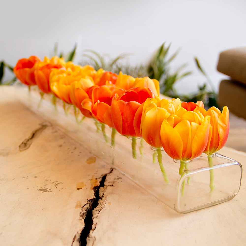 A row of vibrant orange tulips in a long, rectangular clear vase on a light wooden table with a dark natural crack. Green foliage and a beige couch are visible in the background.