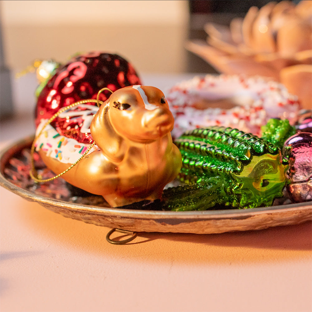 A close-up of a Chive Studio decorative holiday ornament arrangement on a metallic tray, featuring a gold animal ornament, a red strawberry ornament, a green textured ornament